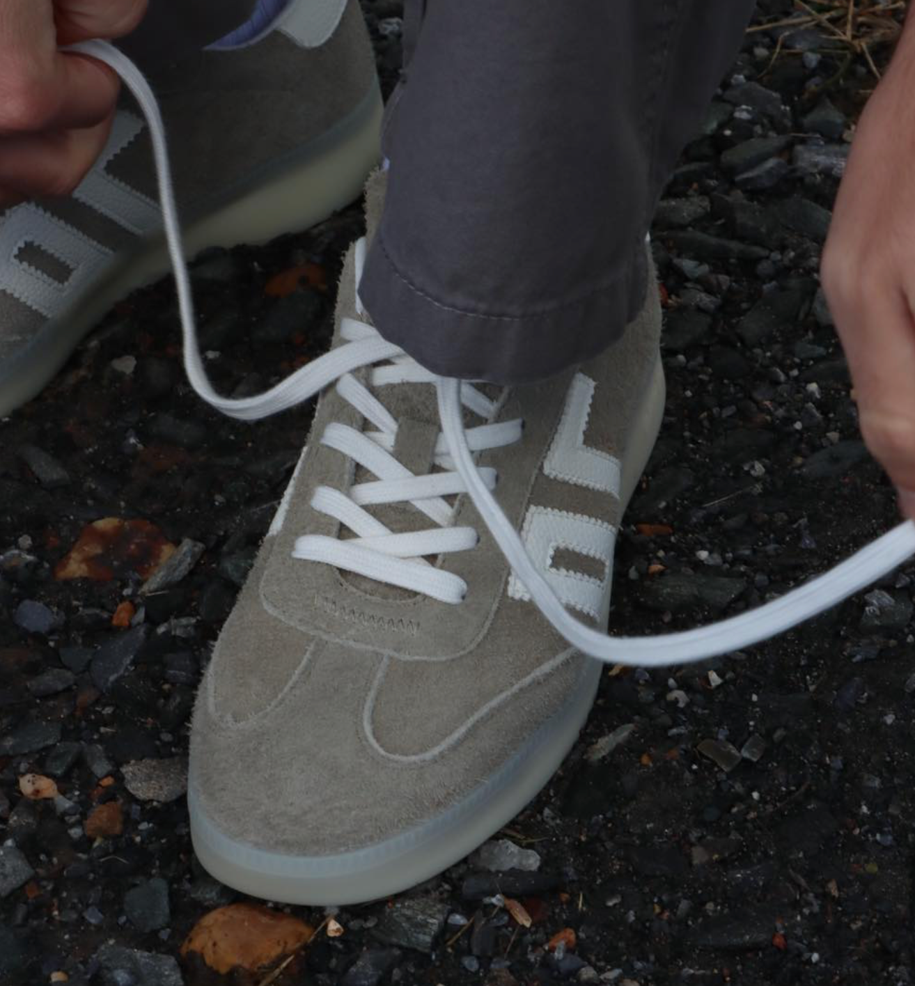 A close-up of a person tying the white laces of a taupe suede sneaker with white side stripes, while wearing dark gray pants, set against a dark gravel background.