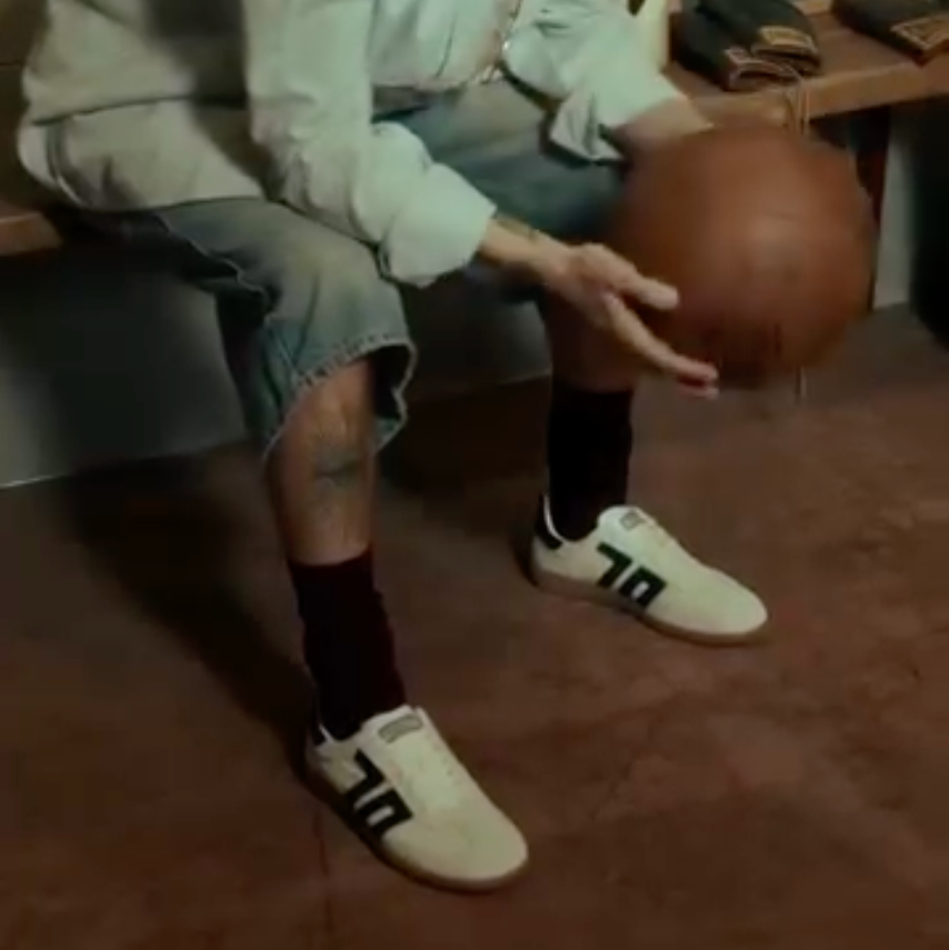 A person with curly hair sits on a bench in a room, holding a basketball. They are wearing a light-colored button-up shirt, denim shorts, black socks, and white sneakers with black stripes.