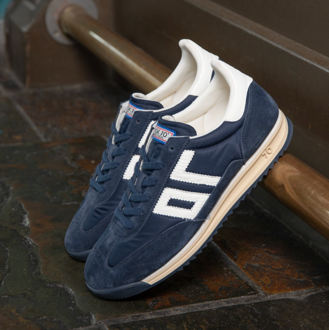 A pair of vintage-style low-top sneakers in navy blue suede and white fabric, featuring a cream-colored midsole and a small embroidered American flag patch on the tongue, resting on a rustic stone floor next to a wooden railing.