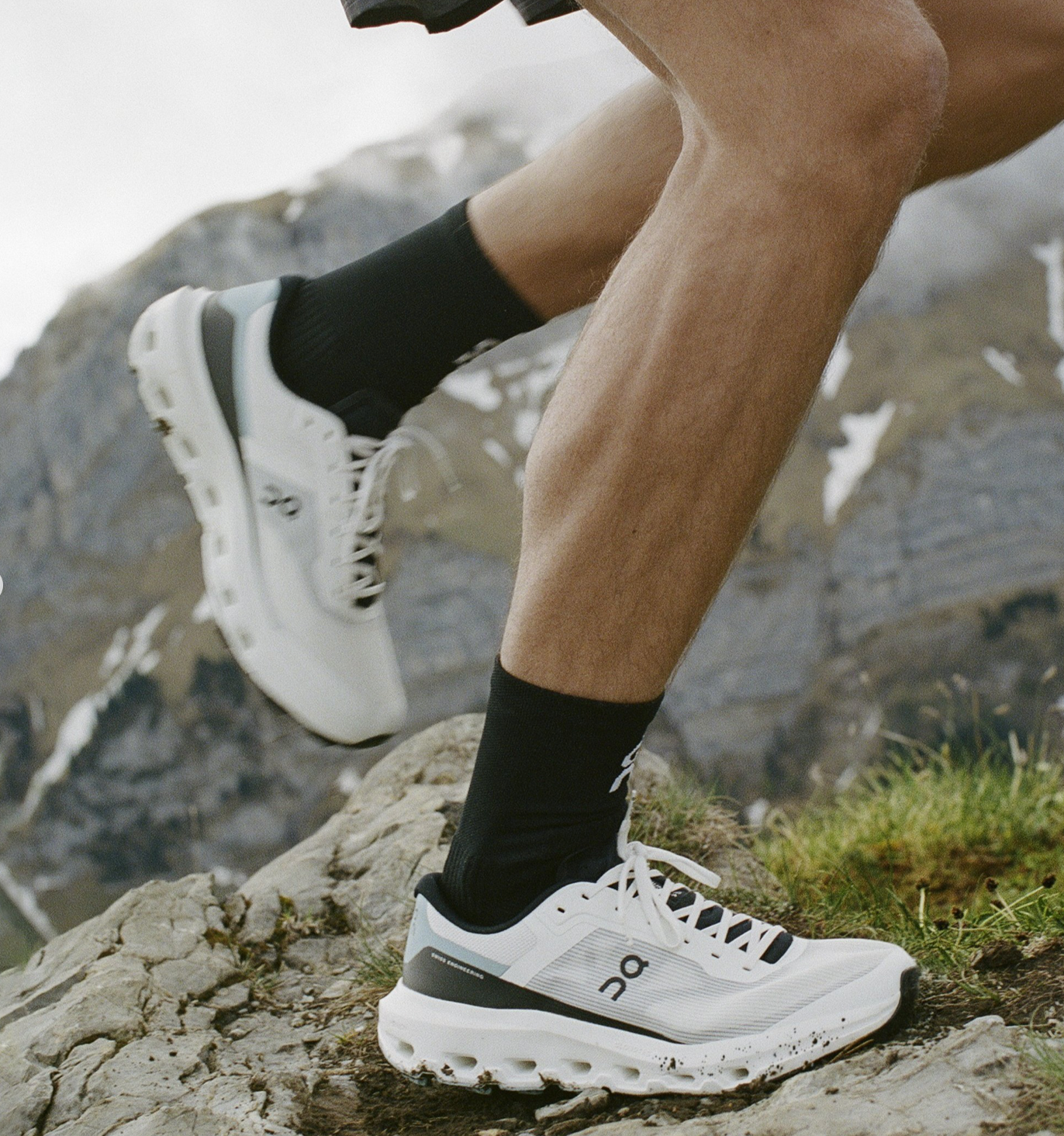 A woman trail running through rocky, snowy mountains, wearing a light purple t-shirt, black shorts with a subtle pattern, a gray headband, and white trail shoes.