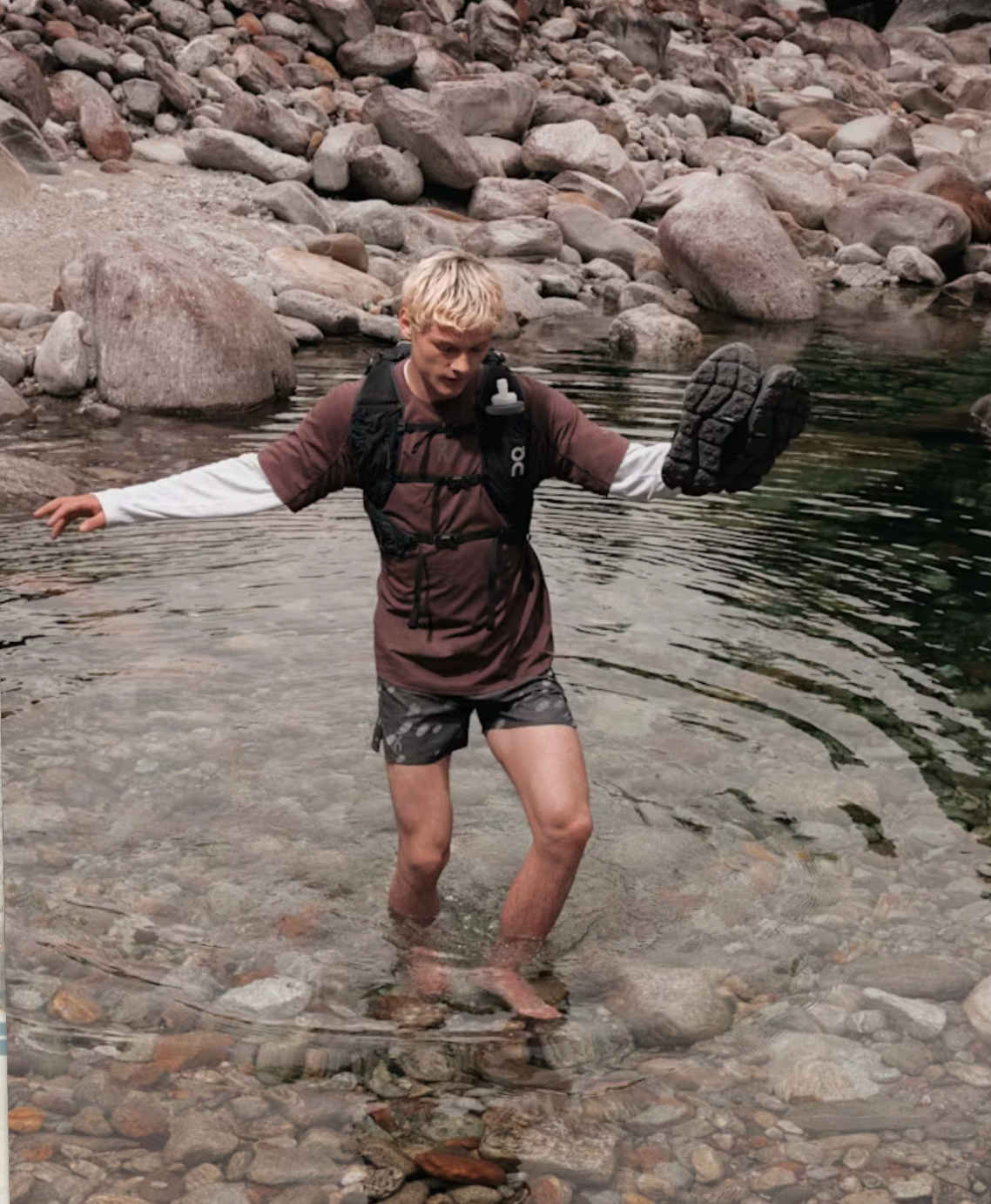 A man standing in shallow rocky water holds up a black boot while wearing a brown and black waterproof shell jacket over a white long-sleeve shirt, with a backpack visible.