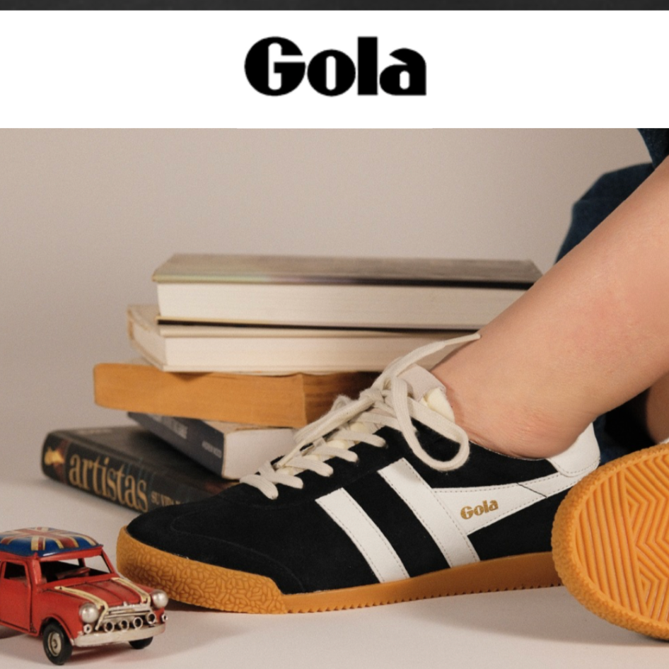A person's foot wearing a black suede sneaker with white side stripes, white laces, and a gum sole, posed next to a stack of books and a small red toy car on a light gray surface.